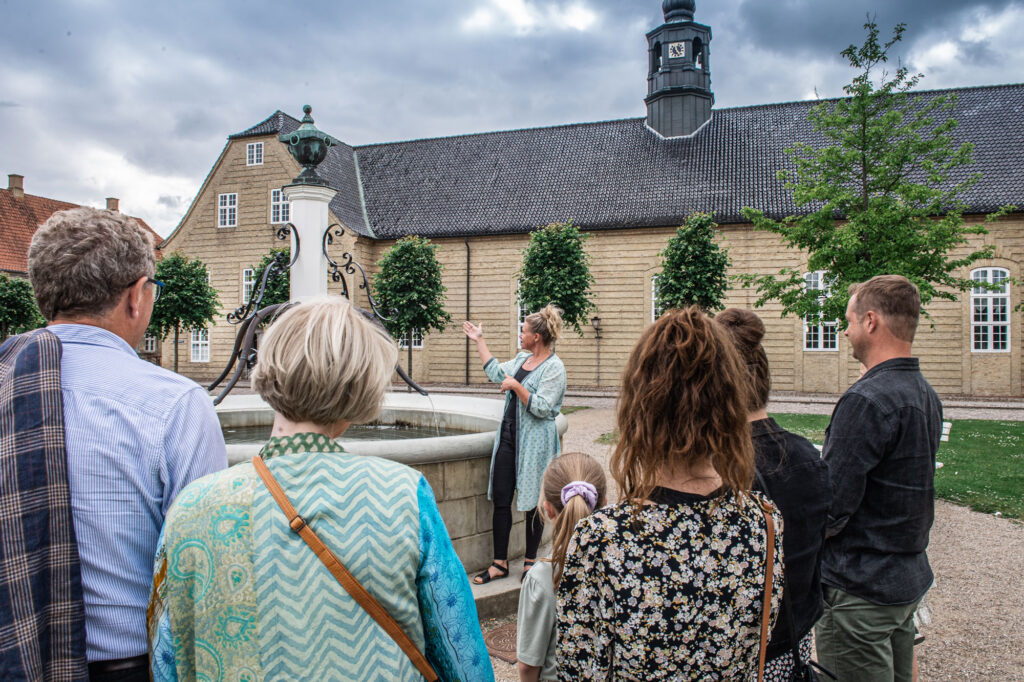 Gruppe på guidet byvandring tur med Museum Kolding i UNESCO byen Christiansfeld på kirkepladsen, hvor guiden fortæller og peger på springvandet mens gruppen lytter og lærer om verdensarv.