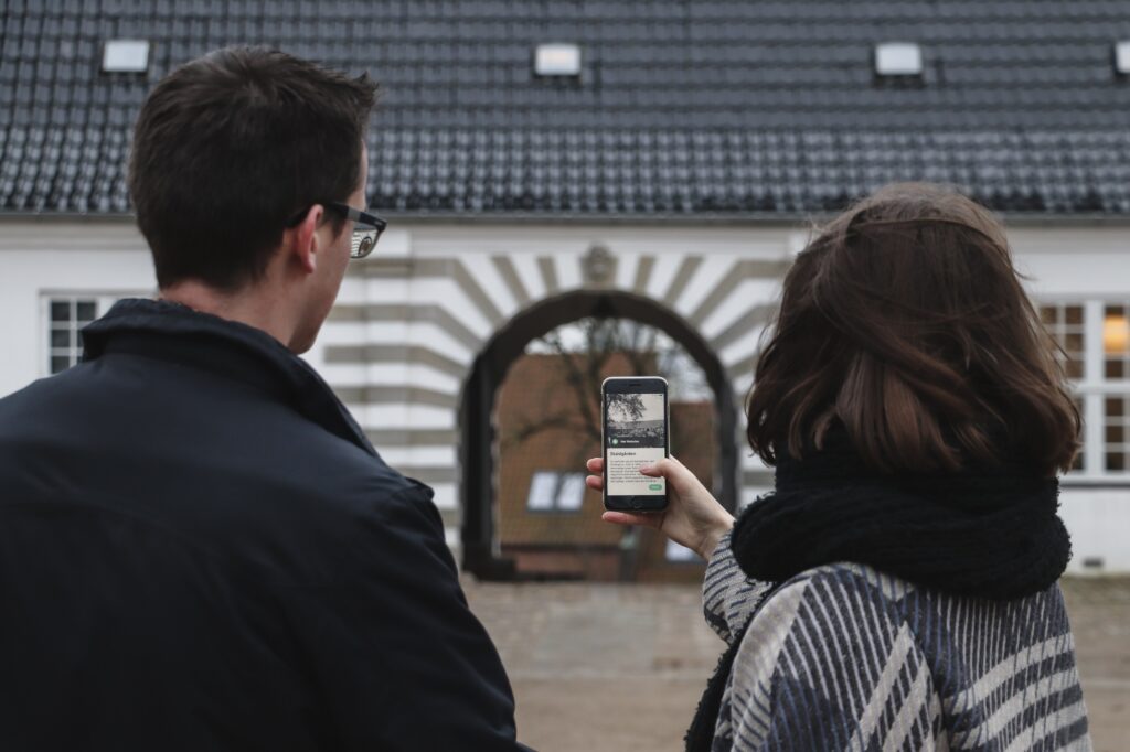 To unge museumsgæster står foran porten til Staldgården og kigger på en smartphone, der viser Vizgu appen med en digital guidet tur på.