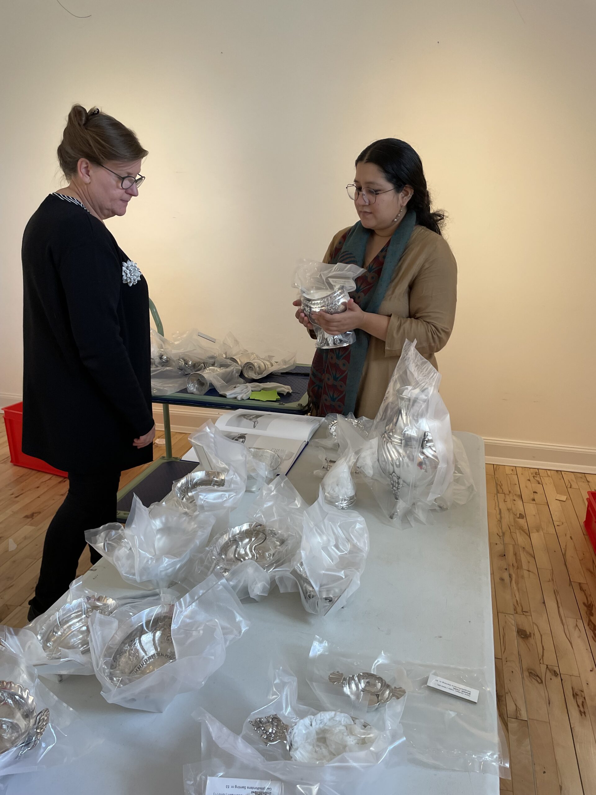 Kurator Komal Pande og museumsinspektør Conni Ramskov står ved et bord omgivet af vakumpakkede sølvobjekter.