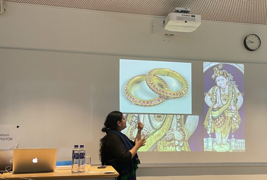 Kurator Komal Pande afholder forelæsning, en projekter viser guldsmykker og gudesymbolik, Komal peger op på billedet af ringene mens hun fortæller.